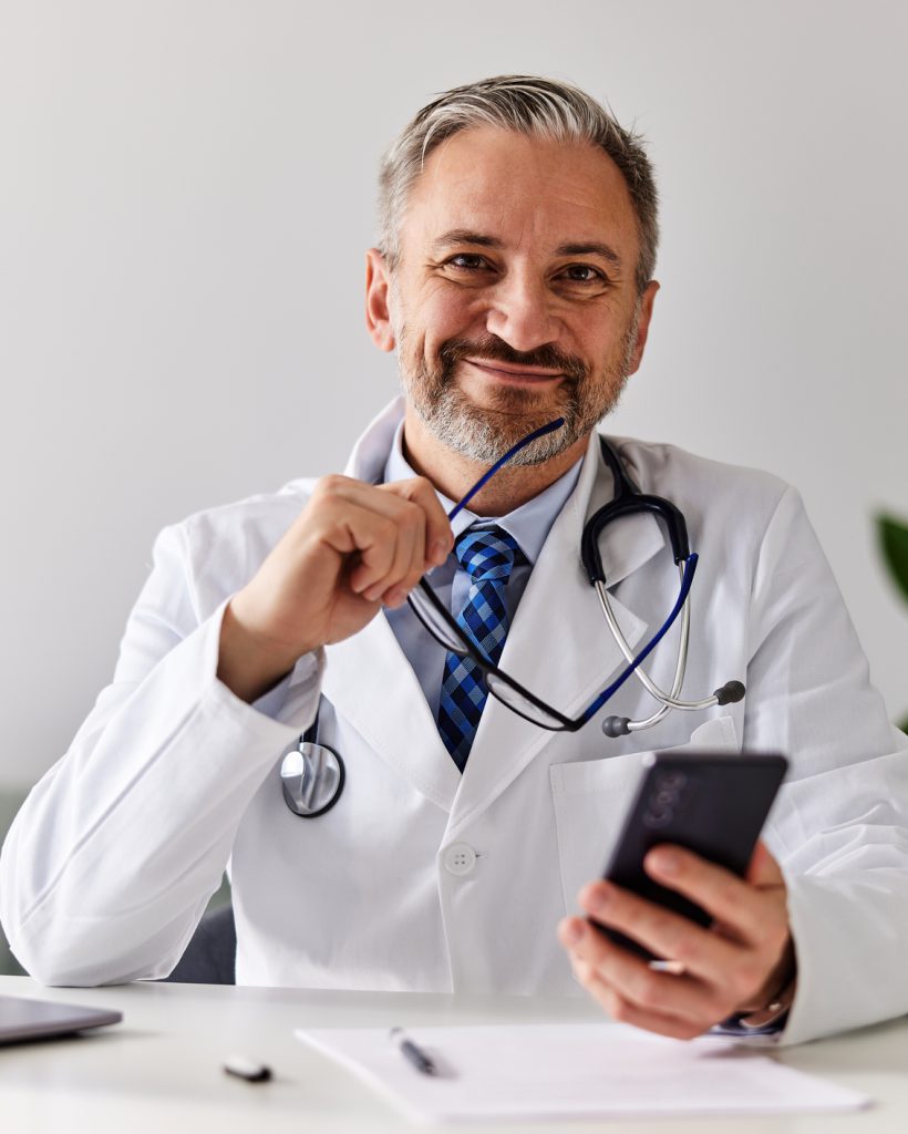 A smiling senior male doctor sitting in front of the laptop, holding a smartphone and gasses, looking at the camera.